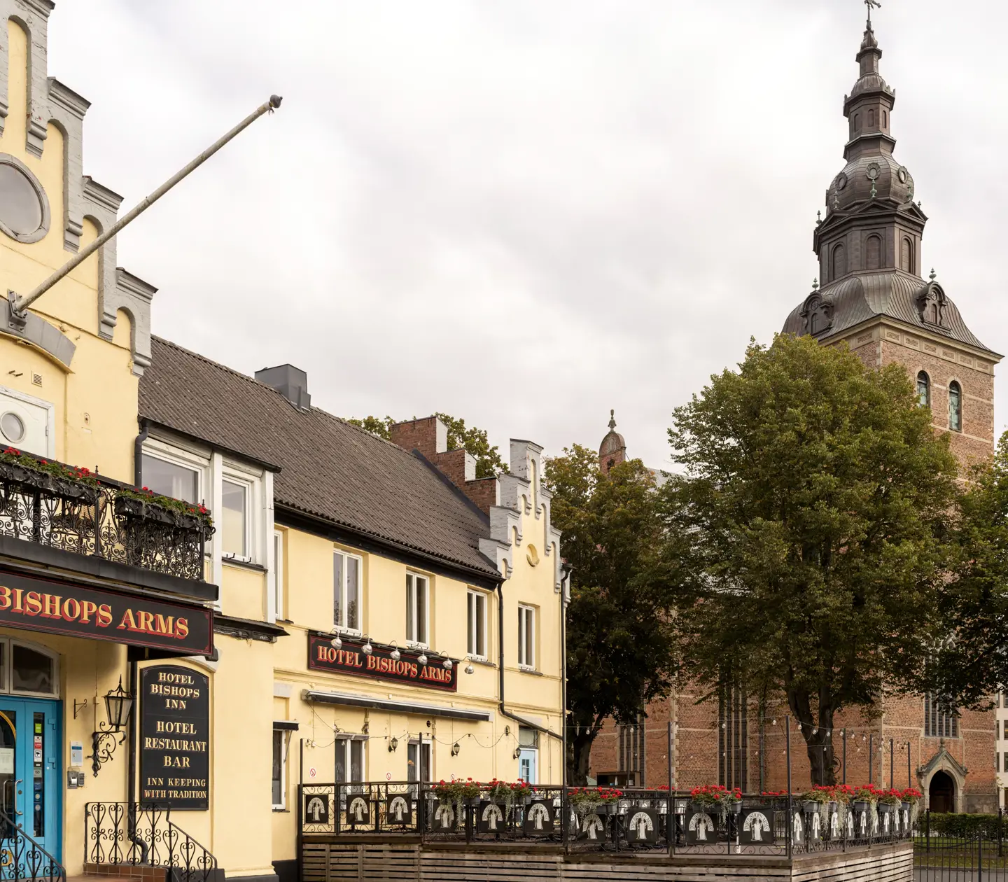 Gula fasaden av Hotel Bishops Arms med kyrka bakom och uteservering framför