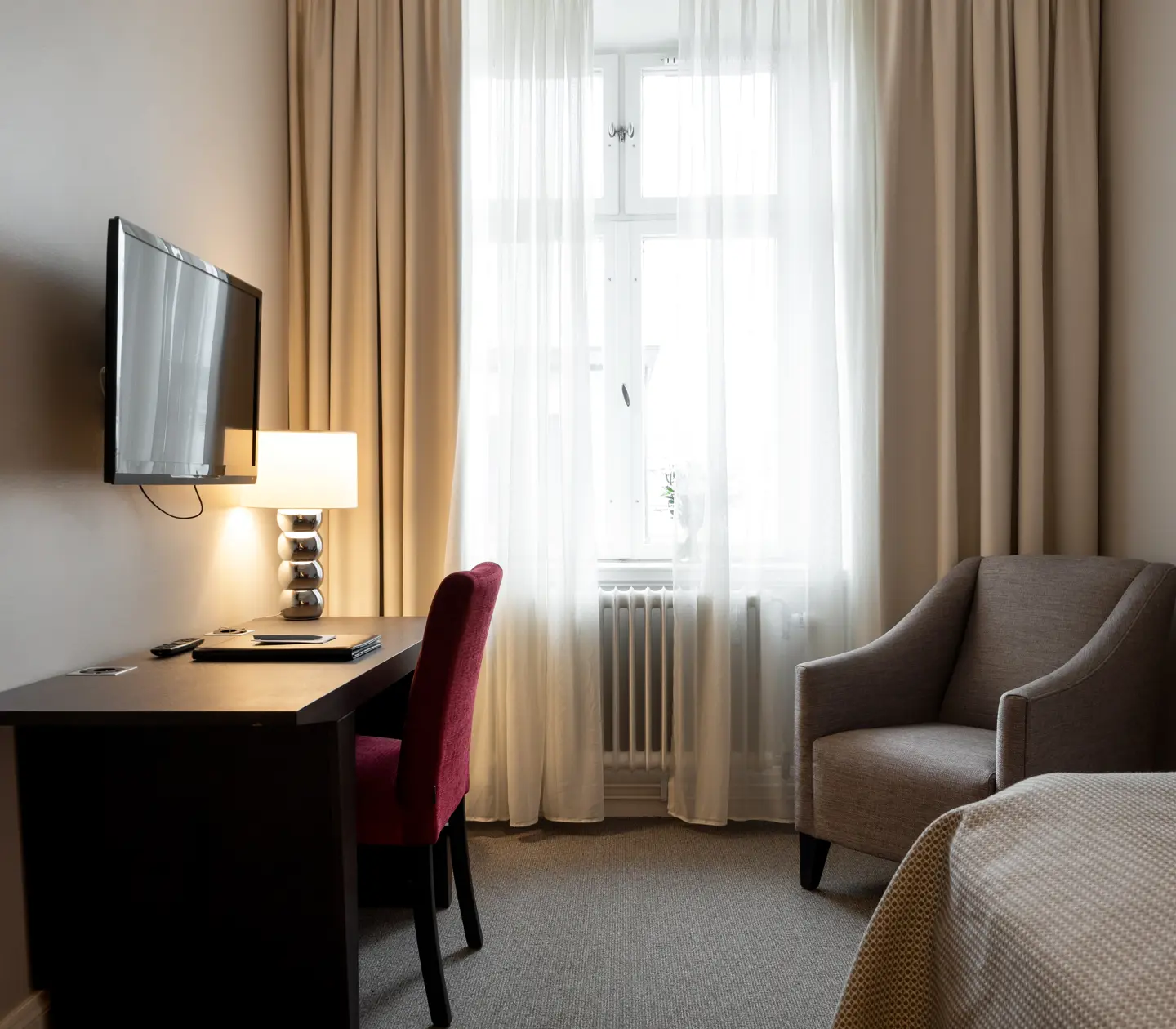Mysigt hotellrum med del av enkelsäng, skrivbord, fåtölj, tv och fönster