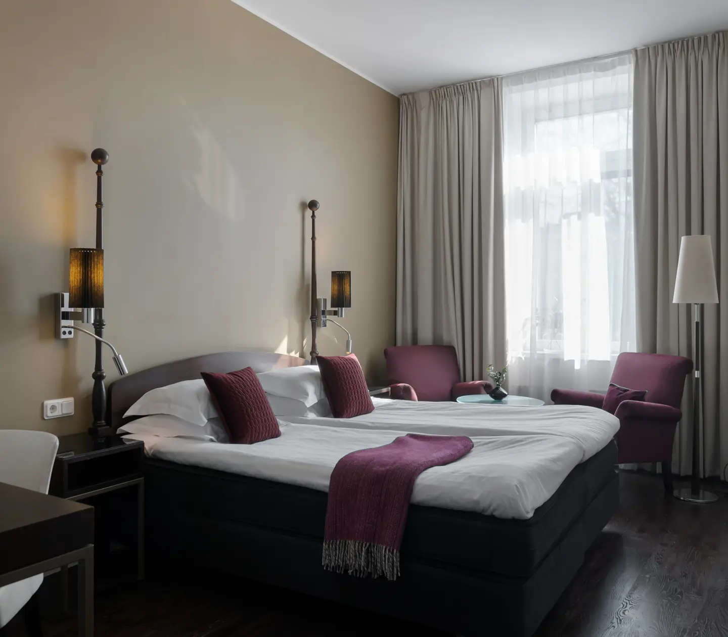 Ljust hotellrum med säng, lila fåtöljer och fönster