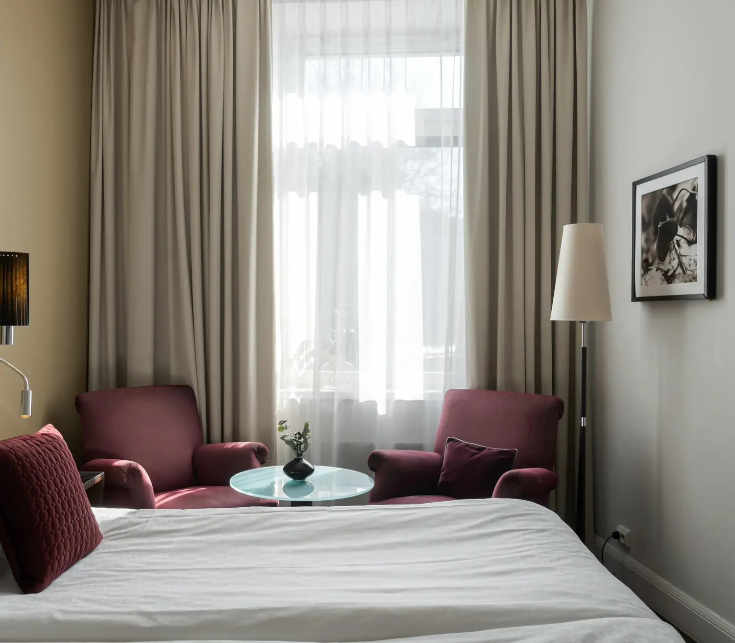 Ljust hotellrum med säng, lila fåtöljer och fönster