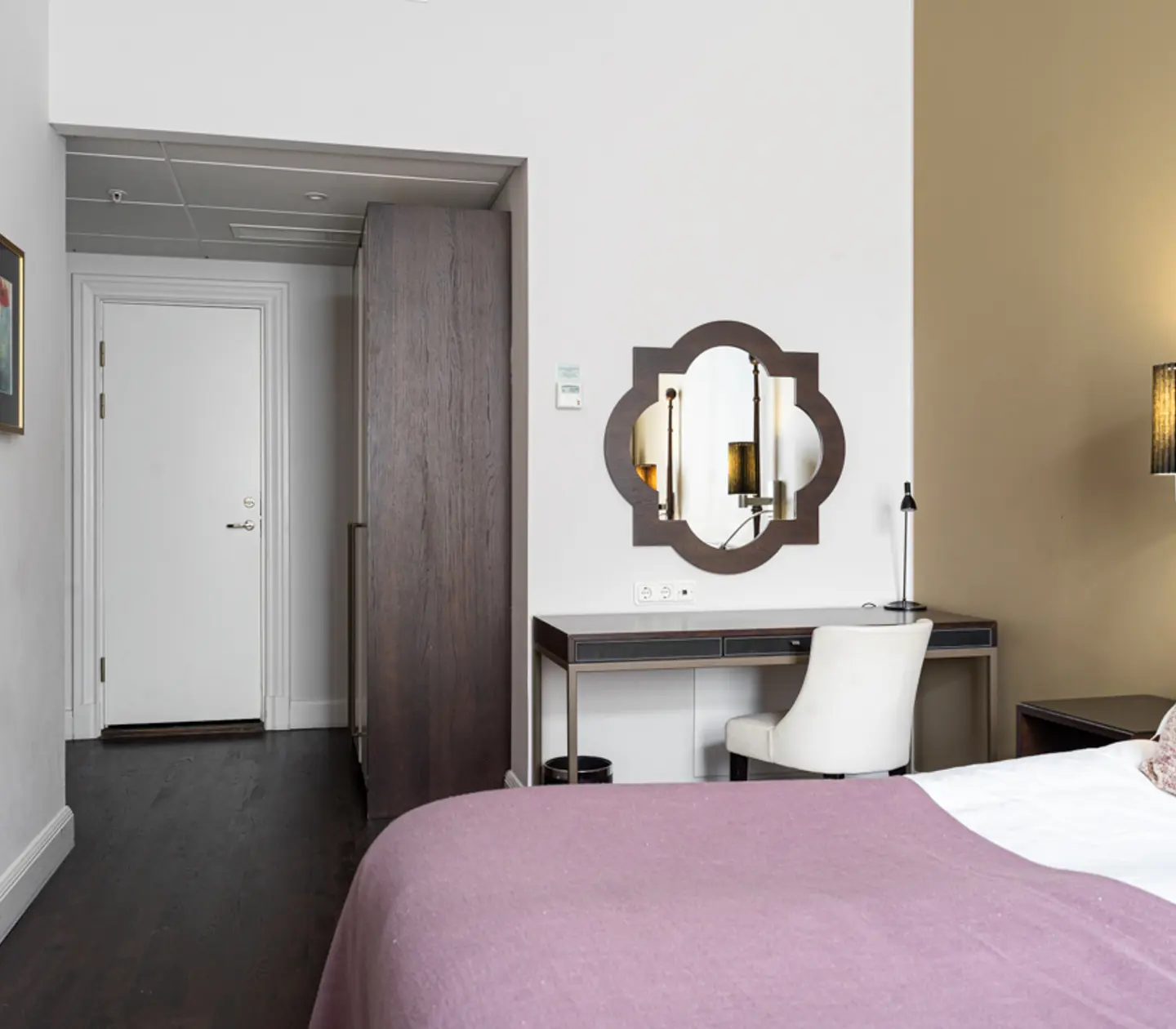 Modernt hotellrum med skrivbord, spegel och säng med lila överkast. Vit stol vid skrivbordet och dekorativa vägglampor.