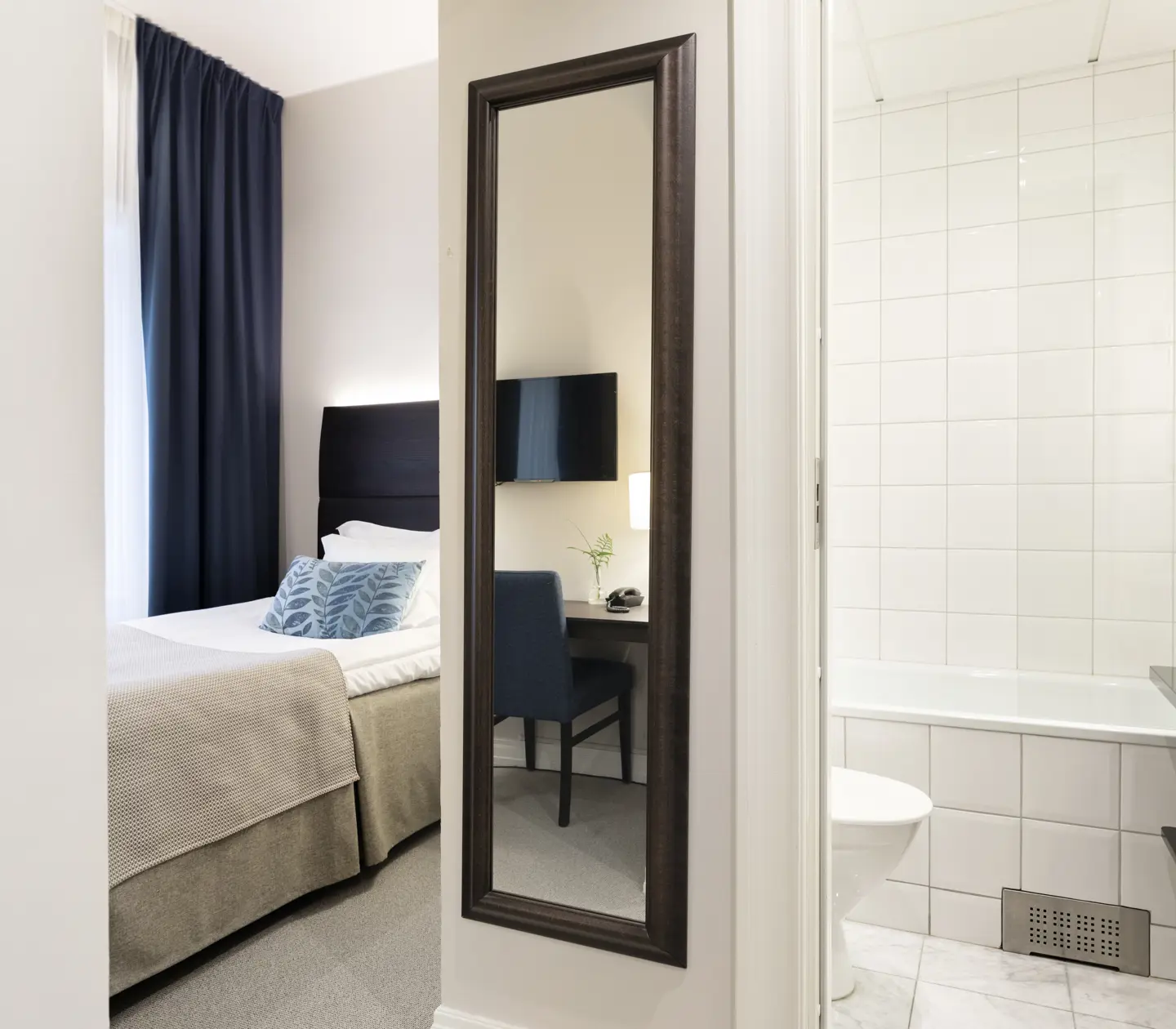 Ljust hotellrum med säng, dörr in till badrum och spegel