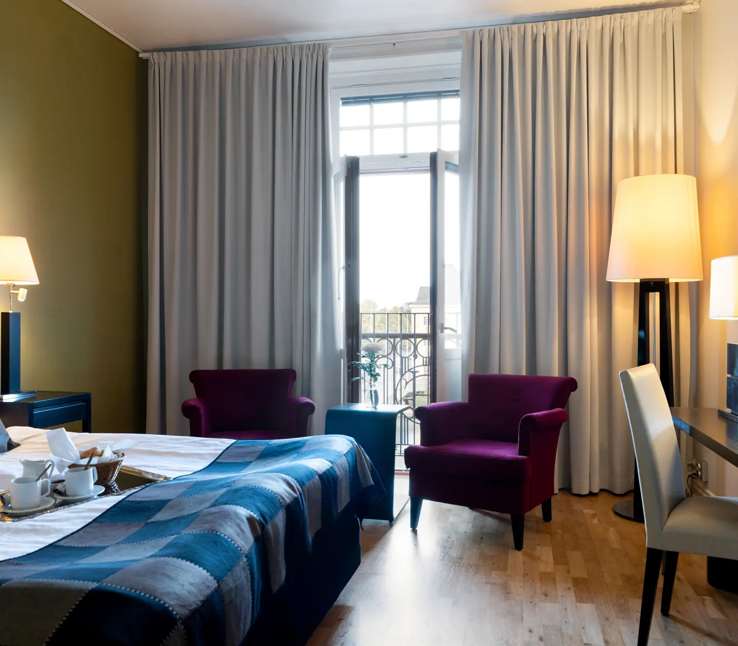 Ljust hotellrum med säng, skrivbord och lila fåtöljer