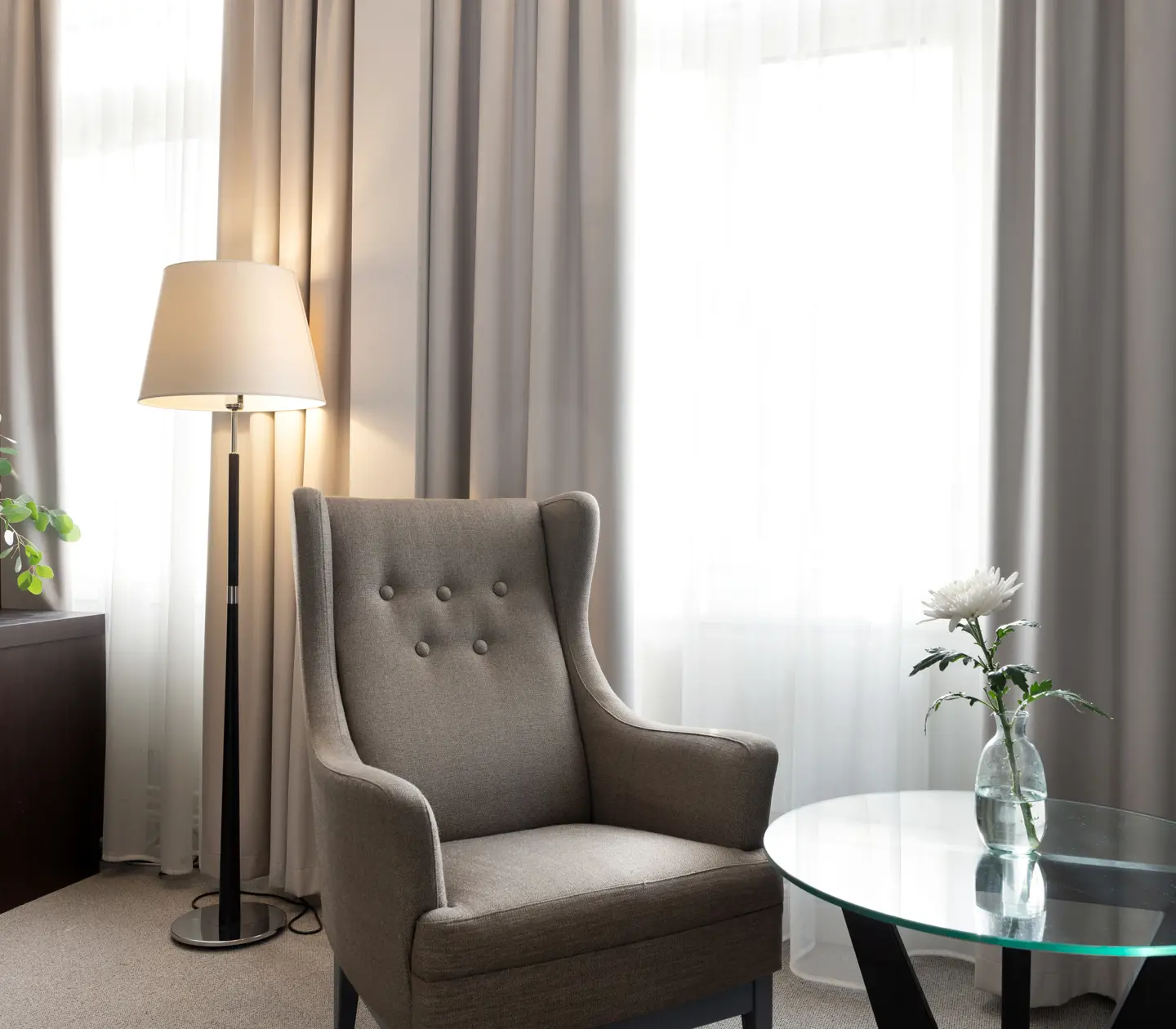 Ljust hotellrum med gardiner, fåtölj och golvlampa