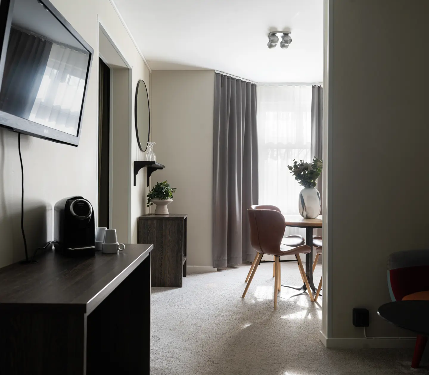 Modernt hotellrum med en plattskärms-TV på väggen och en kaffemaskin på en byrå. I ett ljusfyllt hörn står ett bord med stolar och en vas med blommor. Grå gardiner och en rund spegel kompletterar inredningen.