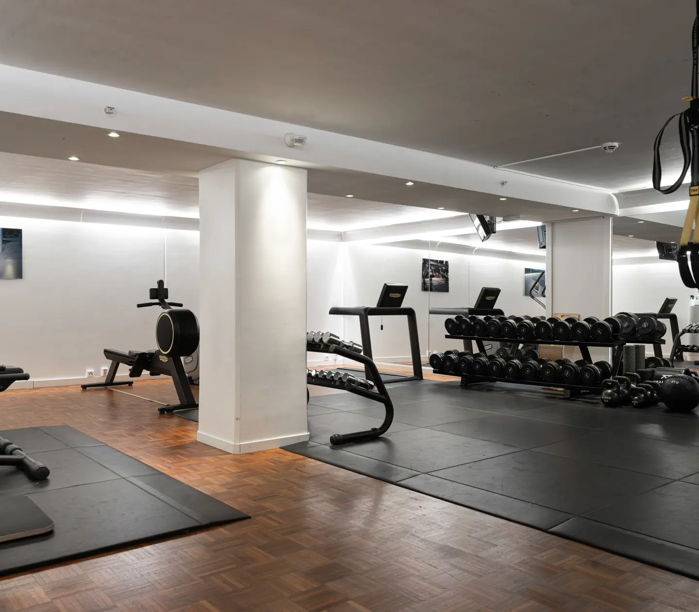 Modern gym med träningsutrustning inklusive löpband, roddmaskin, vikter och TRX-band, i ett välbelyst rum med trägolv och speglar.