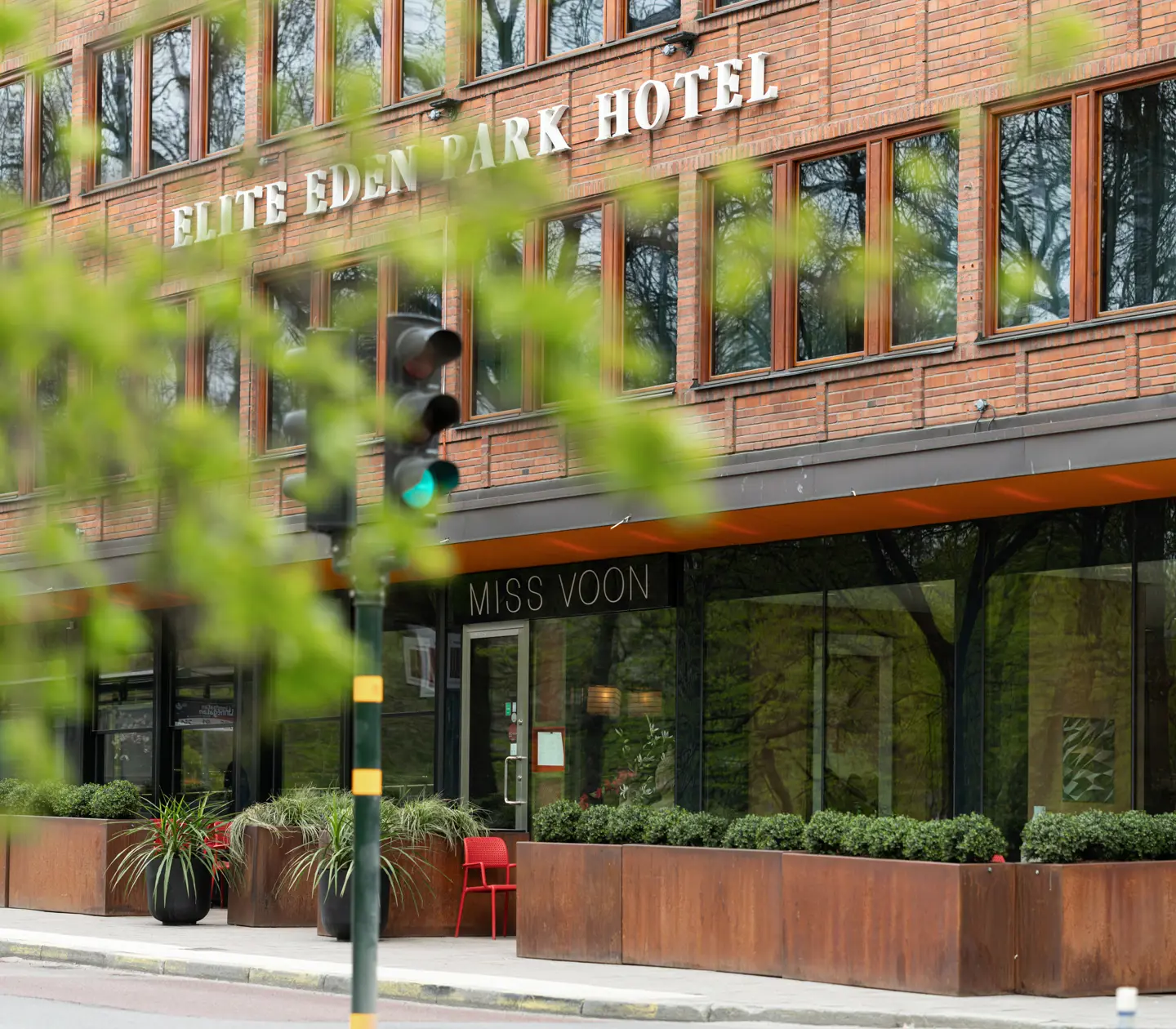 Elite Eden Park Hotel fasad med skylten till restaurang Miss Voon, beläget i en stadsmiljö med grönska i förgrunden.