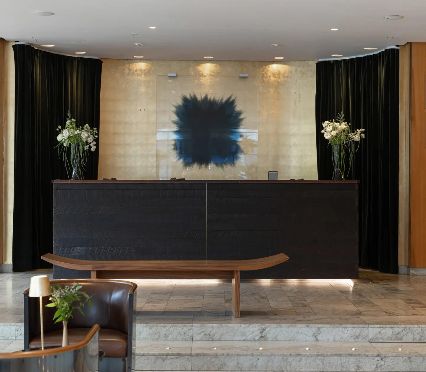 Receptionen på ett modernt hotell med mörkt träskranka, marmorgolv och blomsterarrangemang.