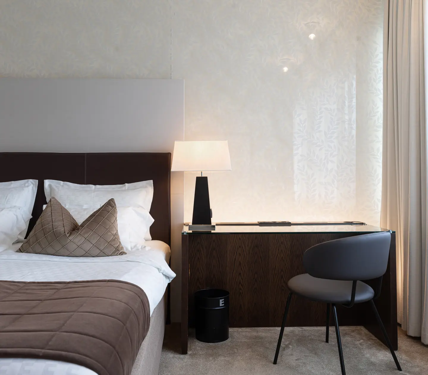 Modernt hotellrum med dubbelsäng, bruna överkast, skrivbord och stol, samt bordslampa vid sidan.