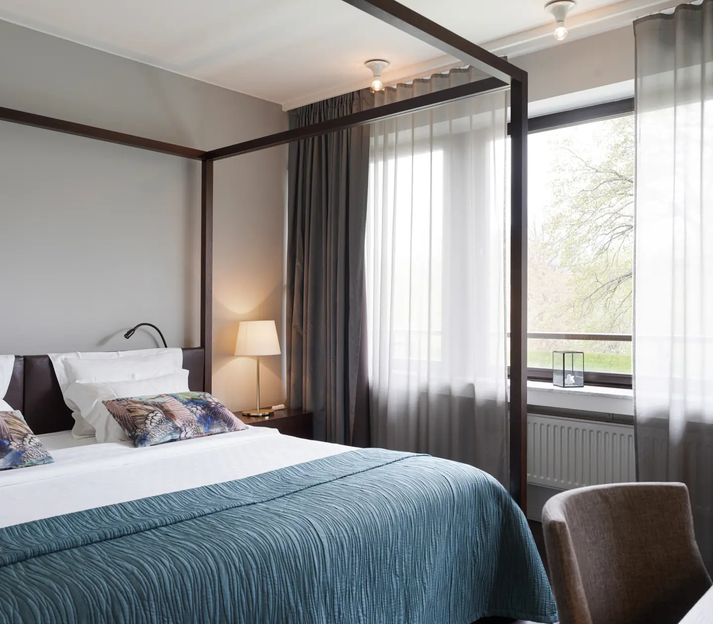 Modernt hotellrum med himmelssäng, blå överkast och dekorativa kuddar vid stort fönster med ljusinsläpp.