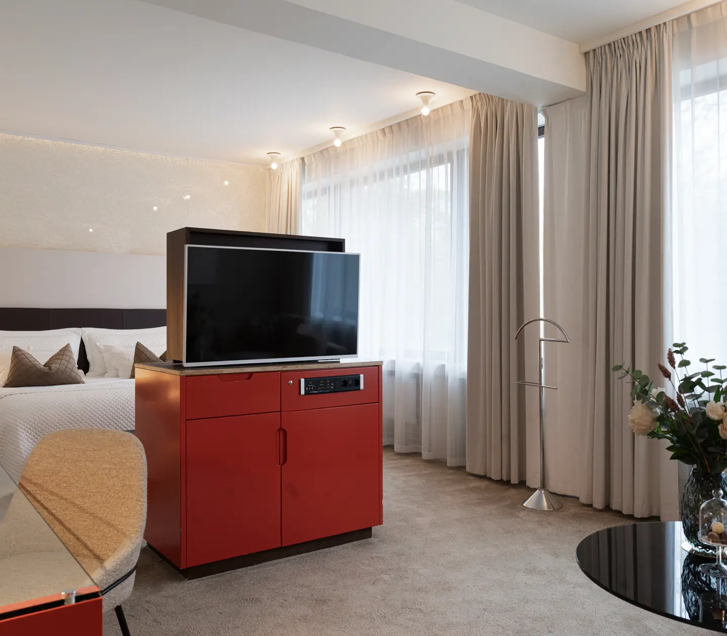 Lyxigt hotellrum med modern inredning, stor TV, bekväm dubbelsäng och blombuketter på ett glasbord. Naturligt ljus strömmar in genom stora fönster med vita gardiner.