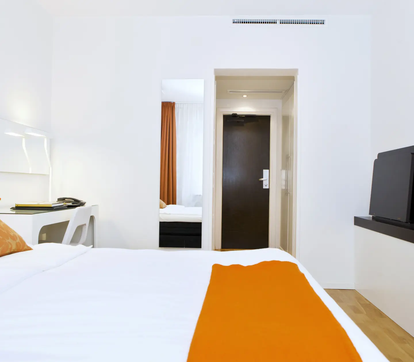 Ljust hotellrum med säng, orange textilier och skrivbord