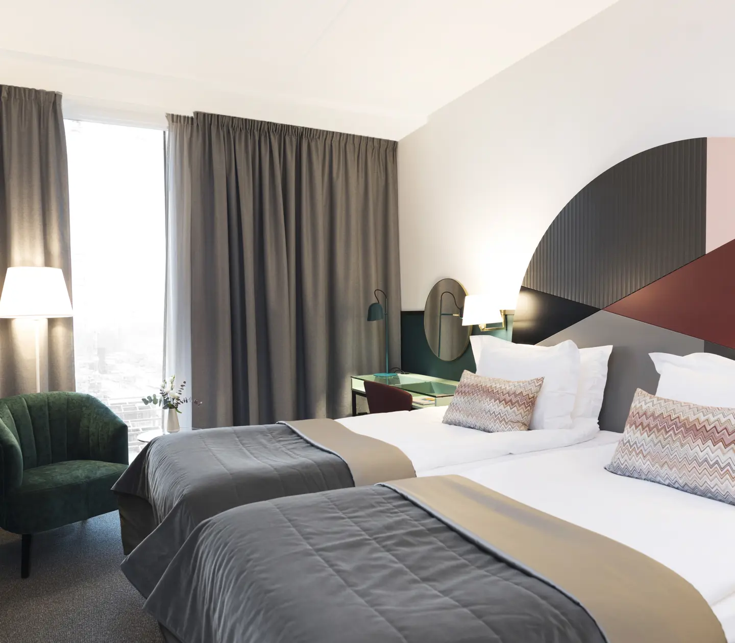 Ljust hotellrum med sängar, färgglad sänggavel och grön fåtölj
