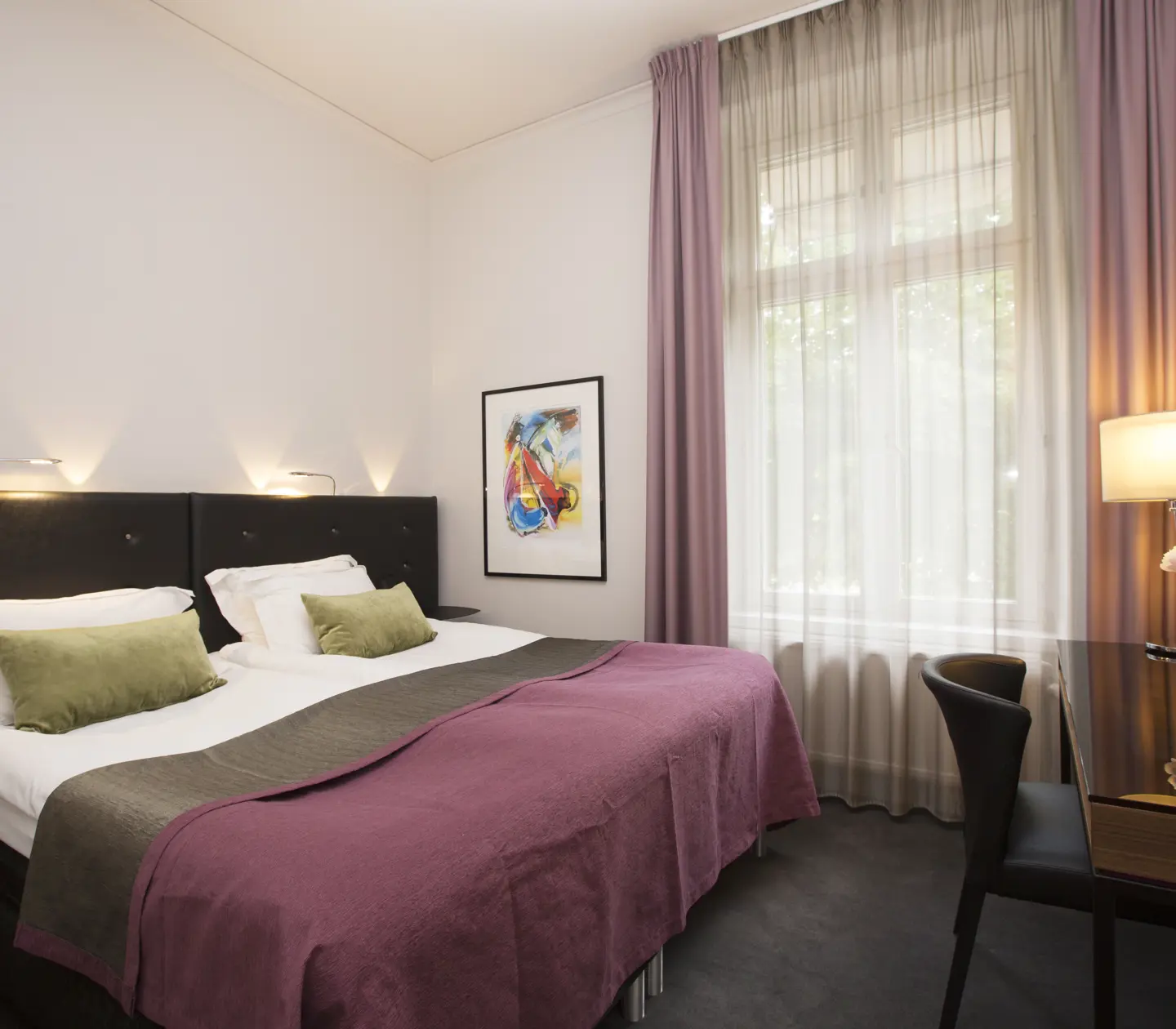 Ljust hotellrum med dubbelsäng, skrivbord, tavla och stort fönster