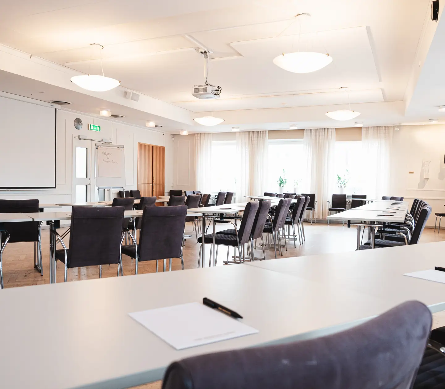 Konferenslokal med många sittplatser, taklampor och projektor