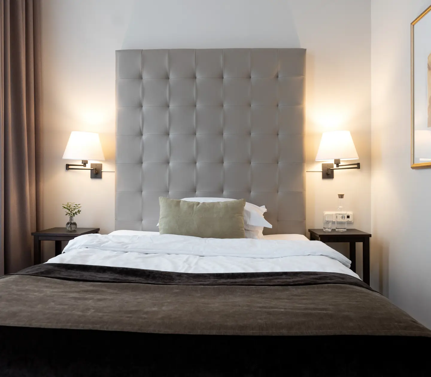 Litet hotellrum med säng, stor grå sänggavel och sänglampor