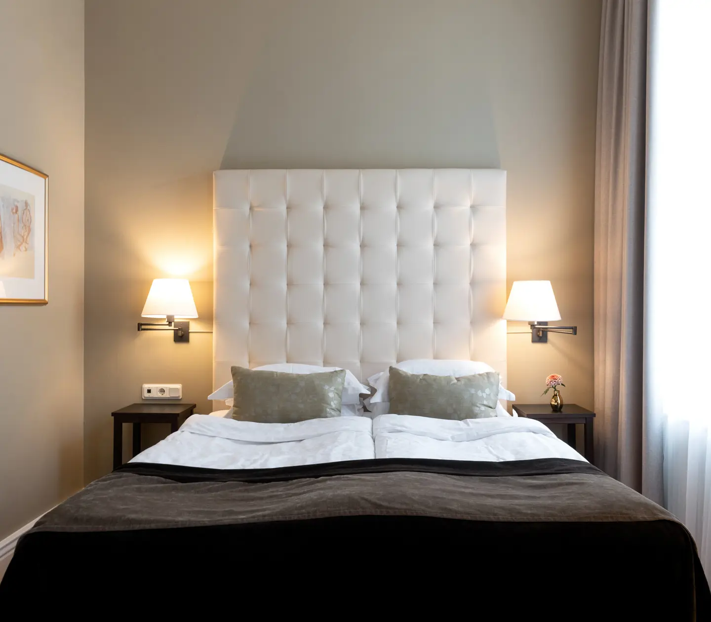 Ljust hotellrum med säng, vit sänggavel och tavla