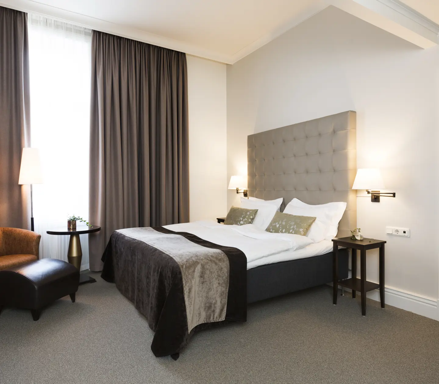 Ljust hotellrum med säng, grå sänggavel och fåtöljer