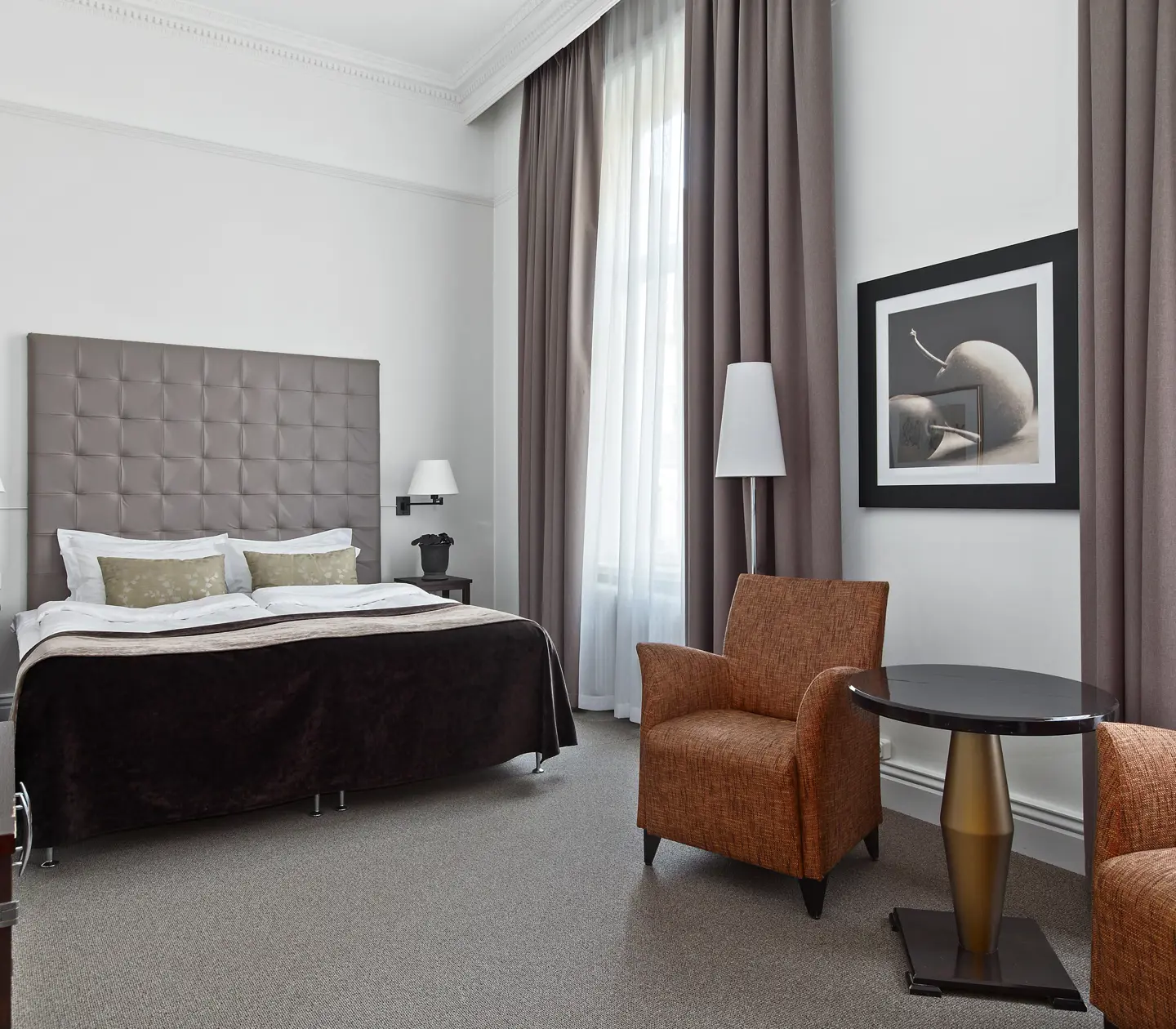 Ljust hotellrum med säng, grå sänggavel och fåtöljer