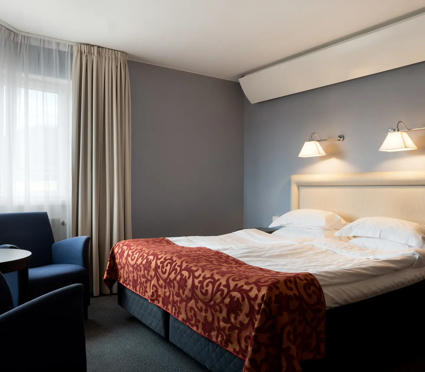Ljust hotellrum med säng, vit sänggavel och fåtöljer