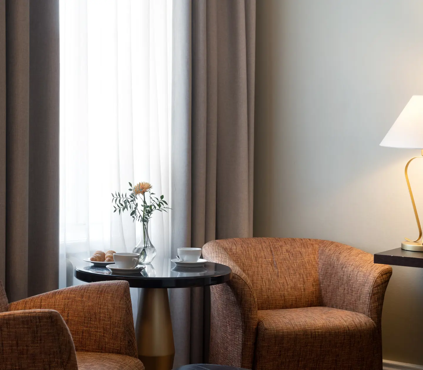 Ljust hotellrum med fåtöljer, bordslampa och kaffekoppar på bord