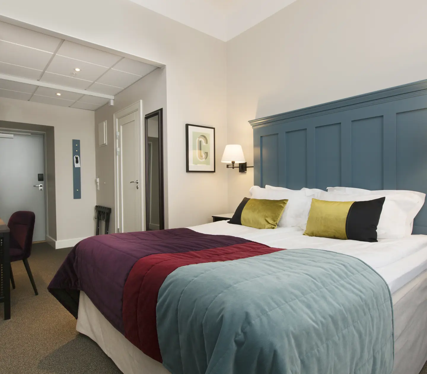 Ljust hotellrum med säng, stor blå sänggavel och skrivbord