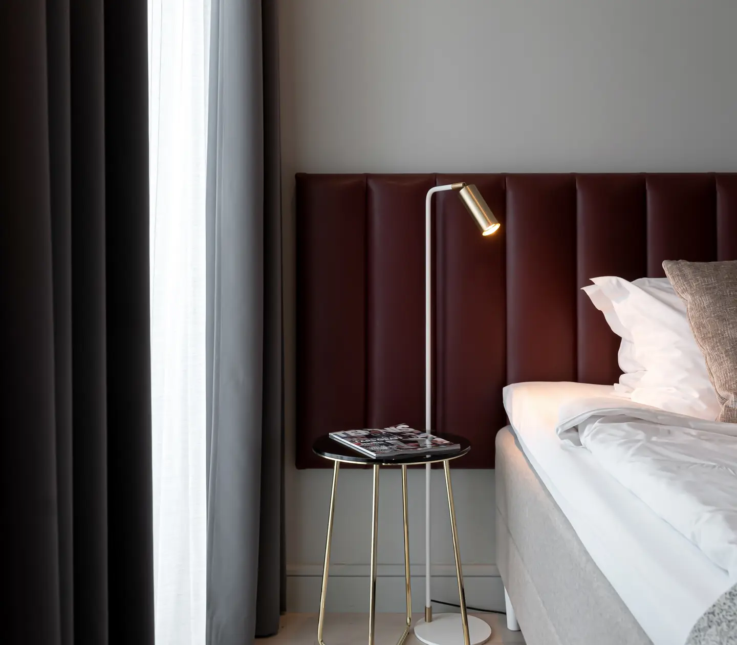 Ljust hotellrum med säng, vinröd sänggavel och liten sänglampa