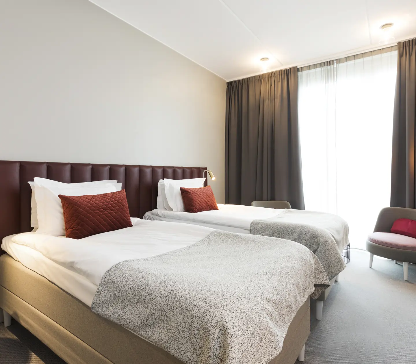 Ljust hotellrum med två enkelsängar bredvid varandra, vinröd sänggavel och fåtöljer