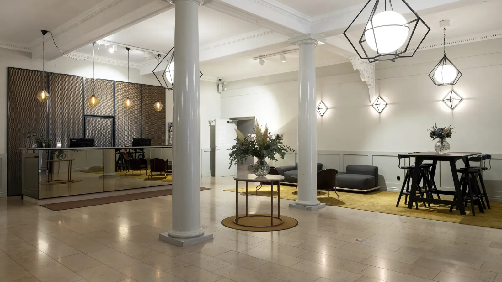 Modern hotellobby med elegant inredning, inklusive en speglad receptionsdisk, stolar och taklampor. Vitmålad pelare och ljus marmorgolv.