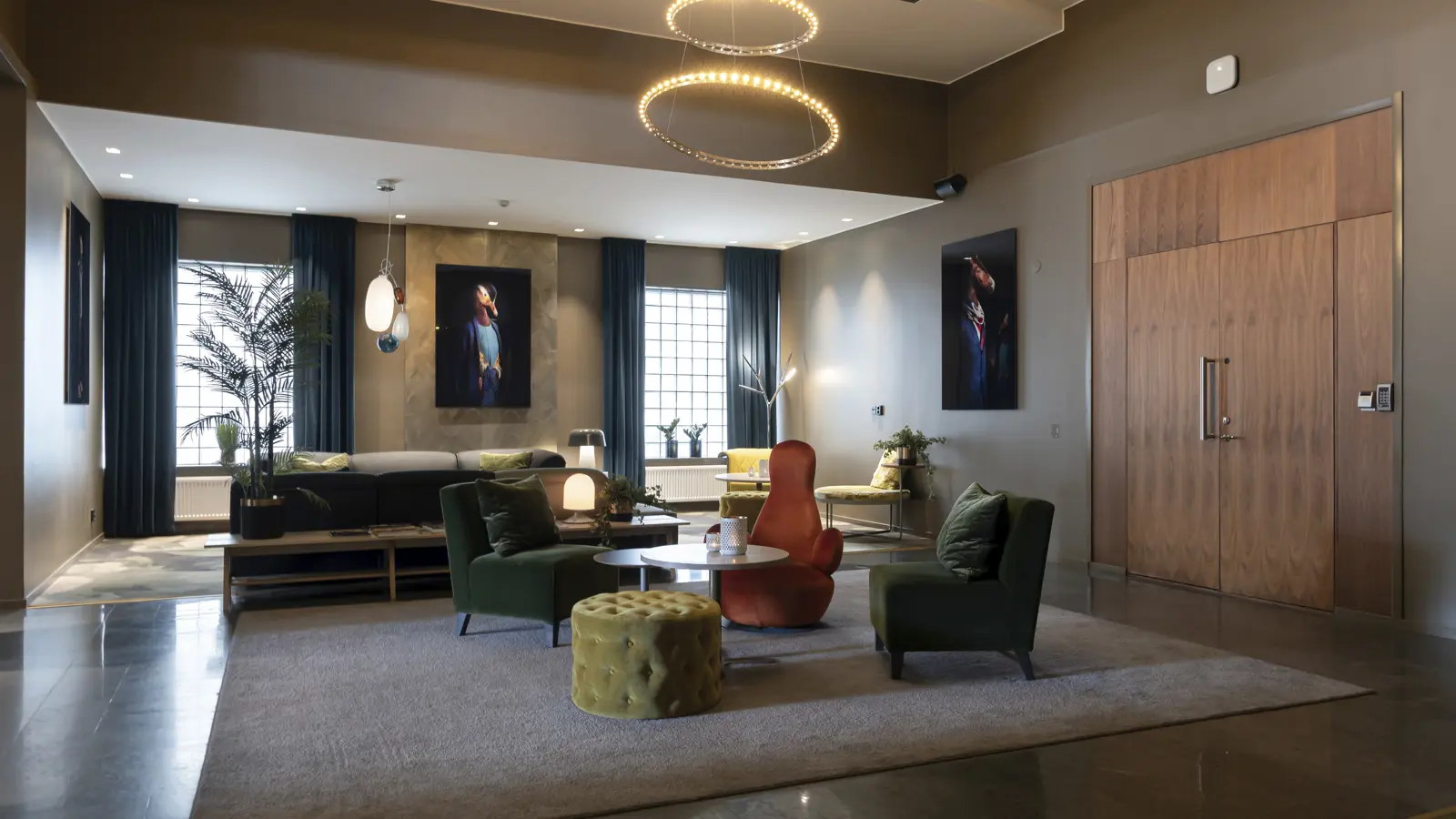 Modern och elegant hotellobby med stilfull inredning, bekväma fåtöljer i grönt och orange, soffgrupp, och konstverk på väggarna. Ljusa fönster, trädetaljer och en cirkulär ljuskrona pryder rummet, vilket ger en varm och inbjudande atmosfär.