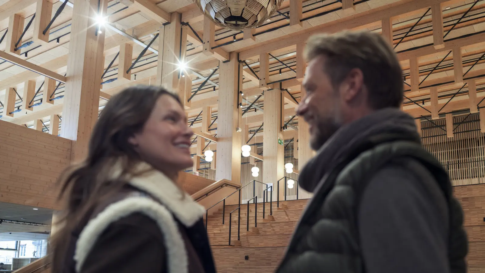 Personer som står inne i en modern byggnad med trädetaljer och stora lampor i taket.