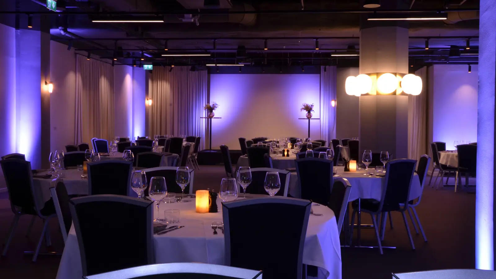 Mörkt upplyst restauranginteriör med runda bord, vita dukar och stolar, dekorerad med glas och levande ljus.