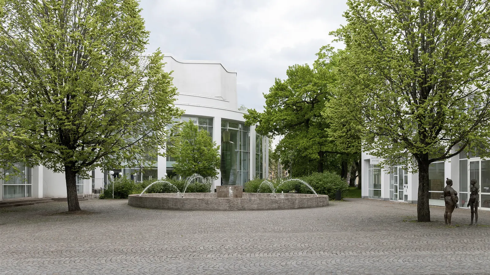 Modern konsthall med träd och fontän i förgrunden, samt skulpturer och grönska omgivna av glasfasader.