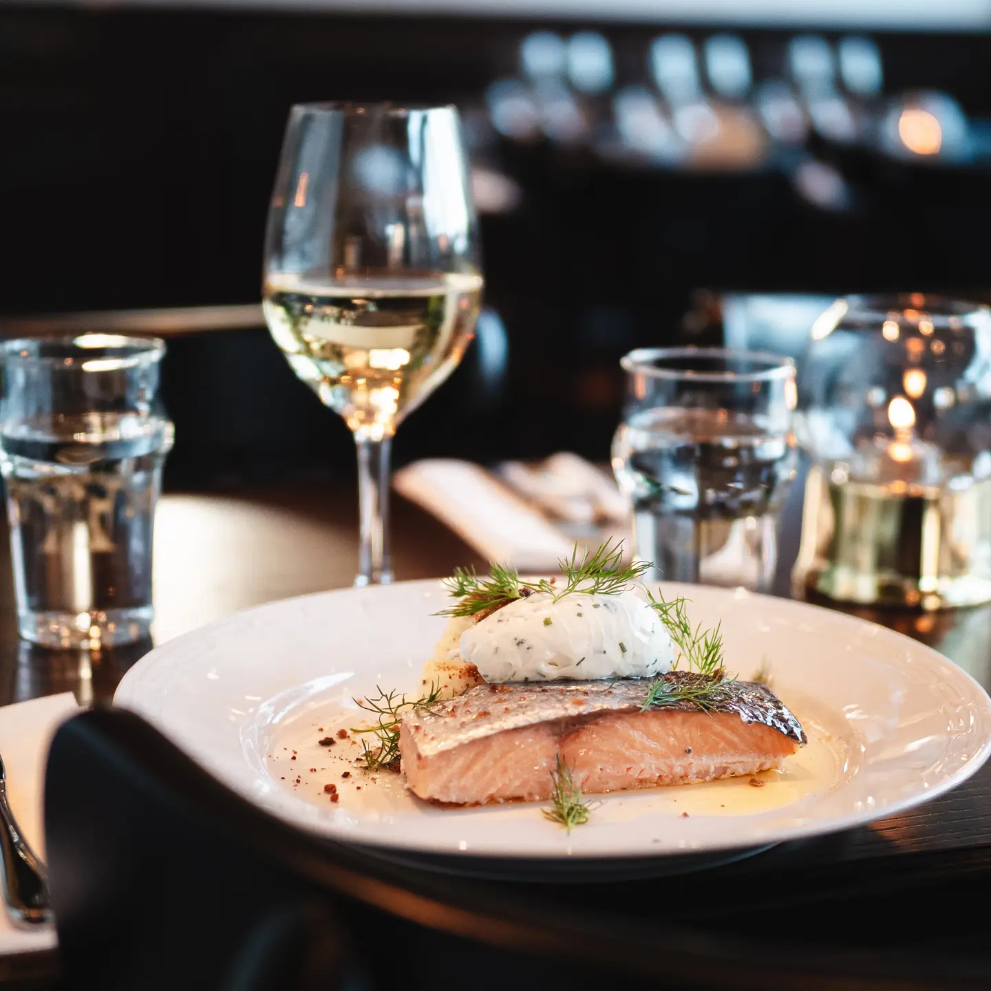 Elegant serverad laxmiddag på en restaurang med ett glas vitt vin, vatten och kristallglas i bakgrunden.